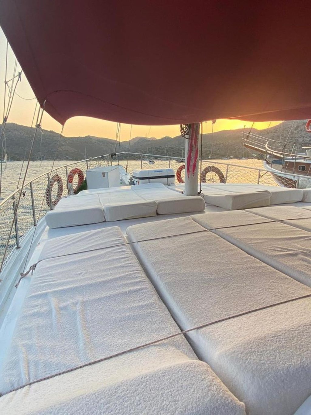 Beyaz minderlerle kaplı yat güvertesi, kırmızı tente ve arka planda deniz ile dağlar.
