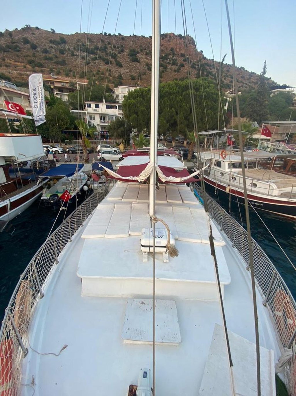 Beyaz güvertesi ve direği olan yelkenli tekne, limanda diğer teknelerin arasında demirli.