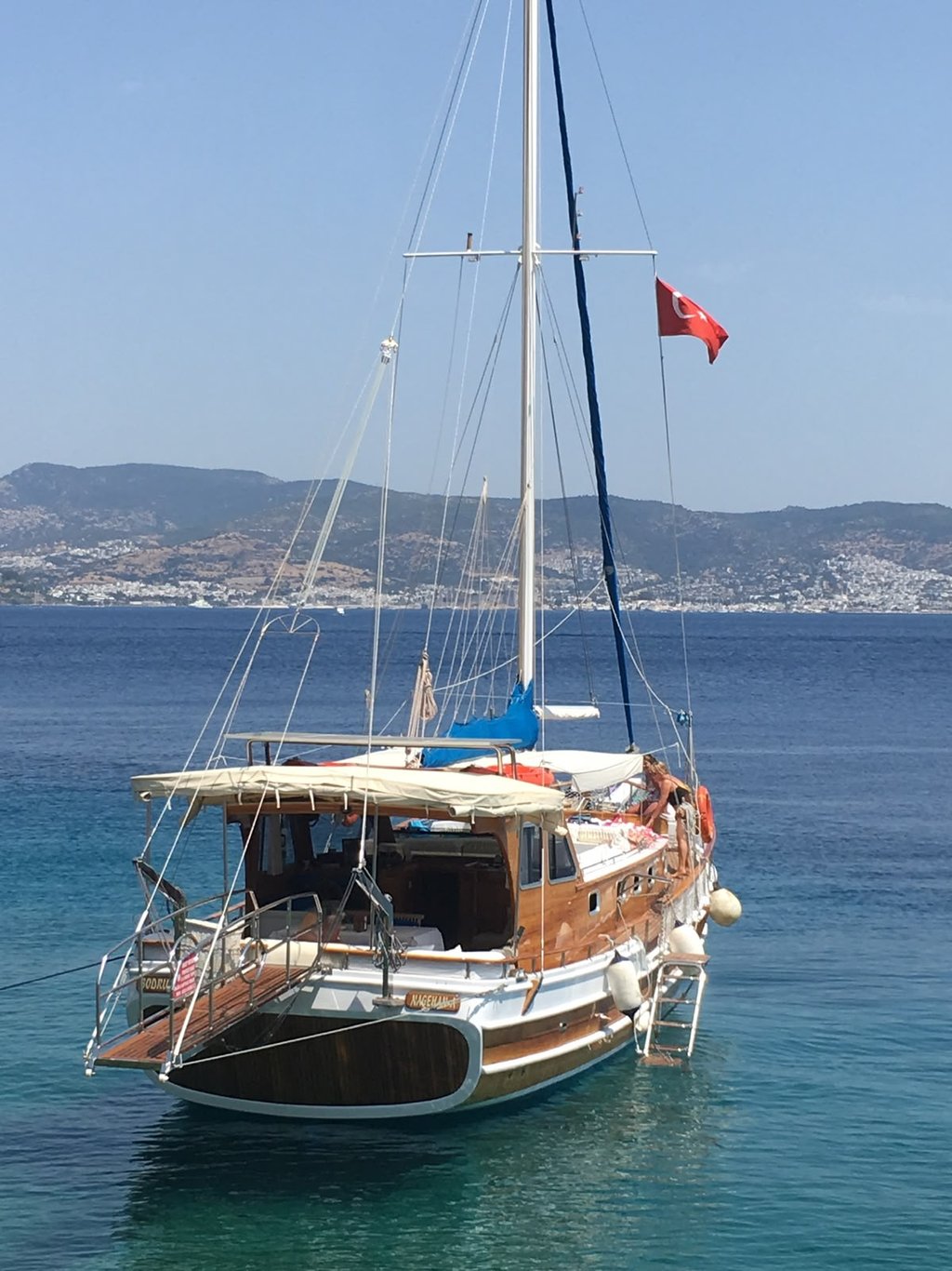 Mavi denizde, Türk bayrağı sallanan ahşap yelkenli tekne ve arka planda dağlar.