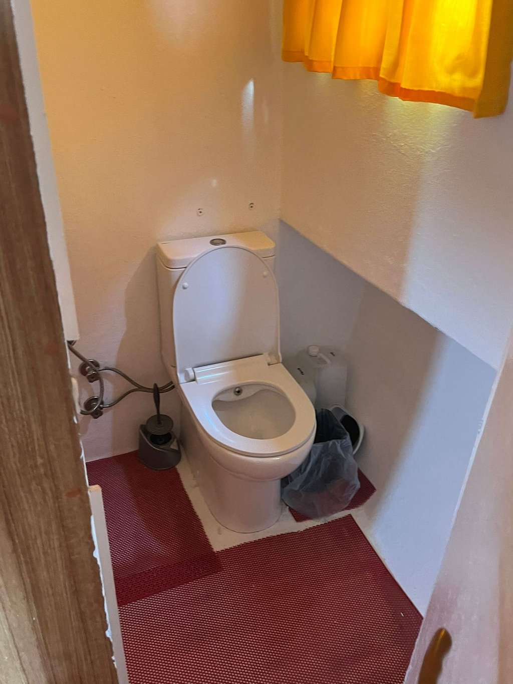 Beyaz klozet, kırmızı halı ve sarı perde bulunan küçük banyo.