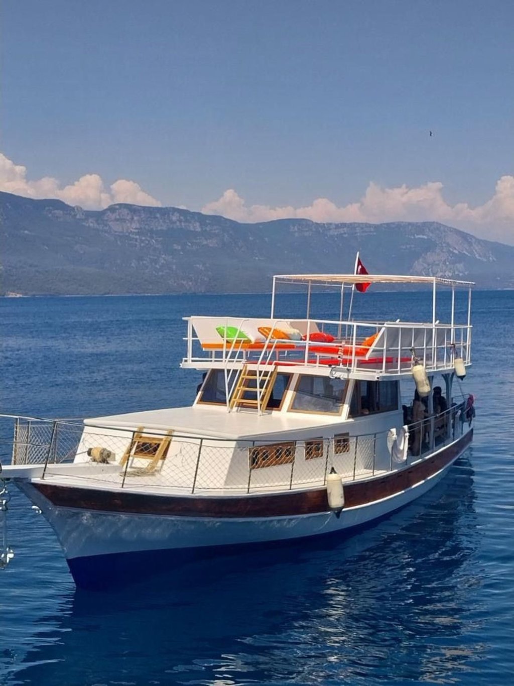 Mavi denizde, üst güvertede renkli minderler ve Türk bayrağı bulunan beyaz ve kahverengi ahşap tekne.