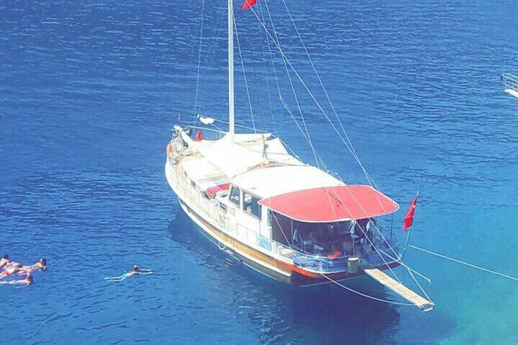 Kırmızı tente ve beyaz gövdesi olan, mavi suda bağlı yelkenli tekne ve çevresinde yüzen insanlar.