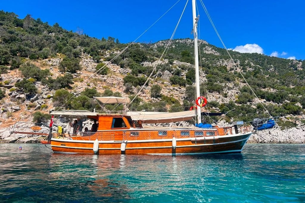 Ahşap gövdeli yelkenli tekne, kıyıda yeşil ve kayalık tepelerin önünde mavi suda.