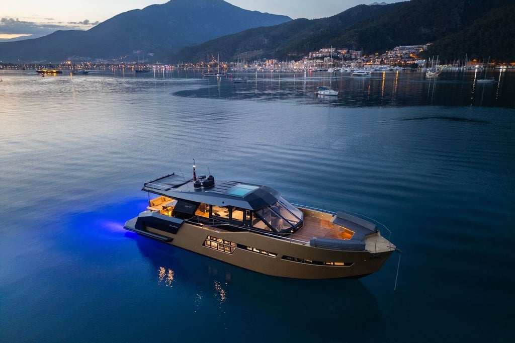 Mavi suda, ışıkları yanan modern motor yat, arka planda dağlar ve sahil kasabası.
