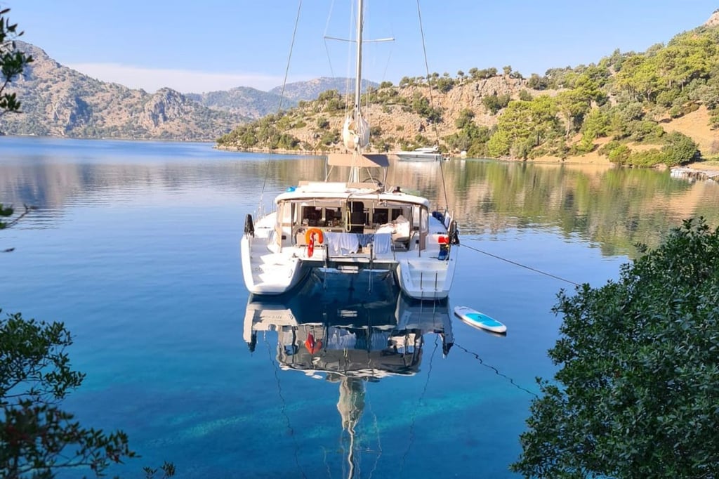 Mavi suda, ağaçlarla çevrili koyda beyaz yelkenli tekne ve küçük mavi şişme bot.