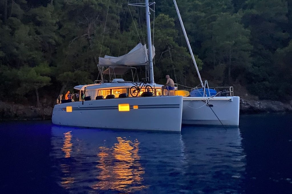 Ağaçların önünde, su üzerinde ışıkları yanan yelkenli katamaran ve güvertede bir kişi.