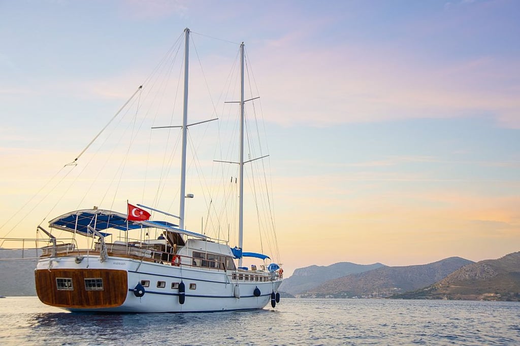 Denizde, Türk bayrağı taşıyan beyaz yelkenli tekne ve arka planda dağlar.