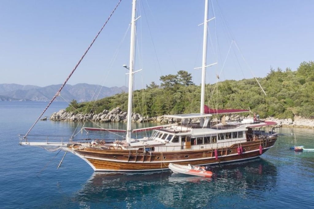 İki direkli, ahşap gövdeli yelkenli tekne, sakin mavi suda, yeşil ağaçların önünde demirli.