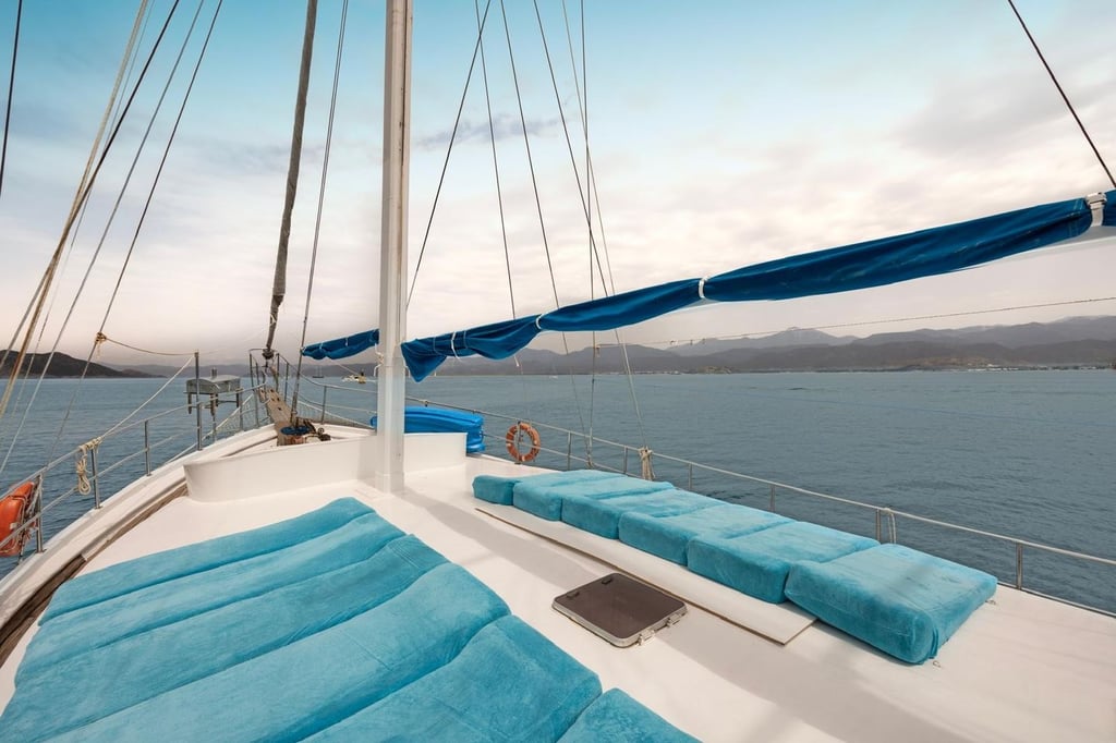 White sailboat with blue cushions on deck and a mast, with sea and mountains in the background.