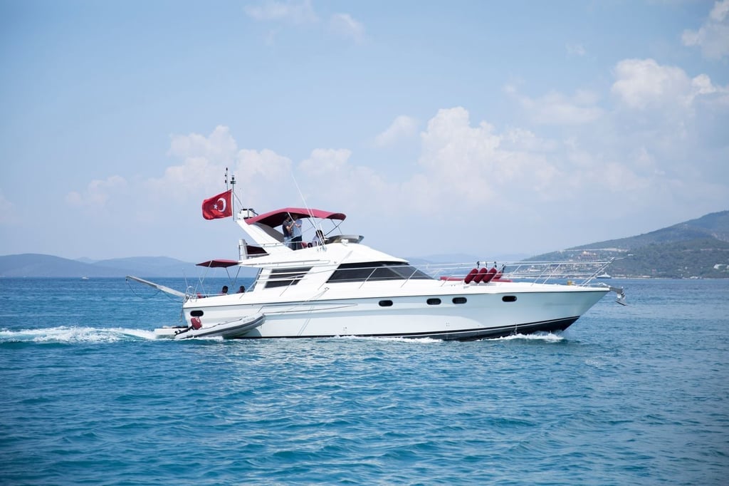 Mavi denizde, kırmızı bayraklı beyaz yat ve arka planda dağlar.