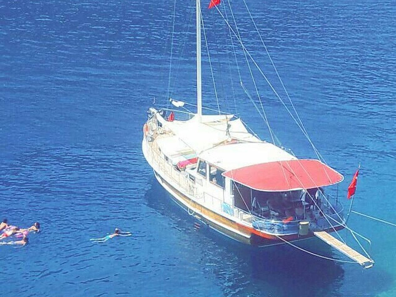 Kırmızı tente ve beyaz gövdesi olan, mavi suda bağlı yelkenli tekne ve çevresinde yüzen insanlar.