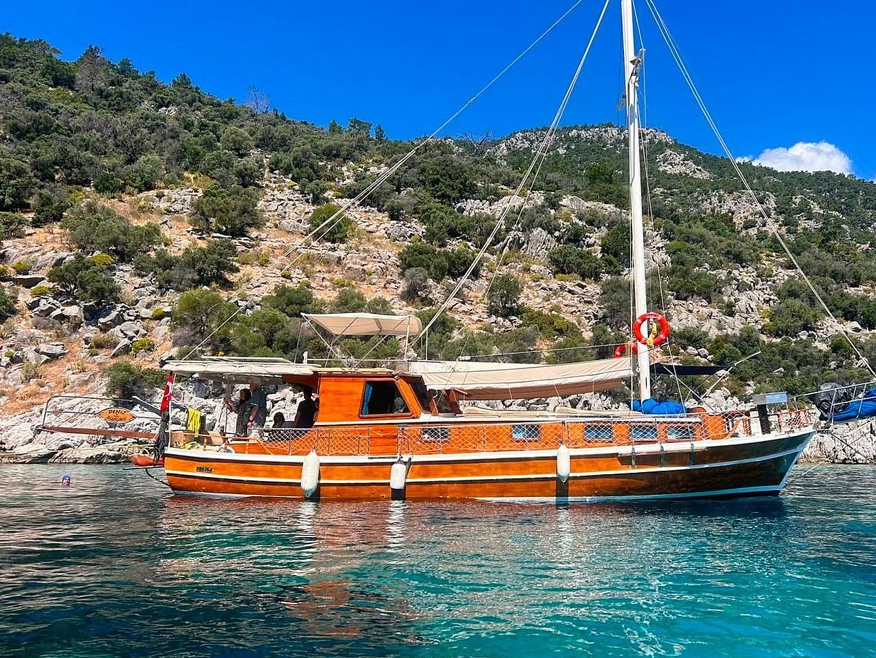 Ahşap gövdeli yelkenli tekne, kıyıda yeşil ve kayalık tepelerin önünde mavi suda.