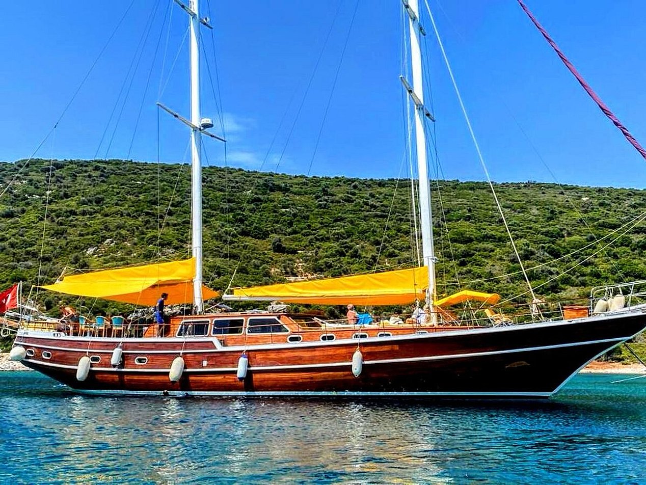 İki direkli, ahşap gövdeli yelkenli tekne, sarı tente ve yeşil tepelerin önünde mavi suda.