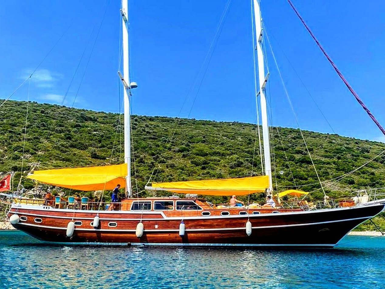 İki direkli, sarı yelkenli, ahşap gövdeli yelkenli tekne, yeşil tepelerin önünde mavi suda.