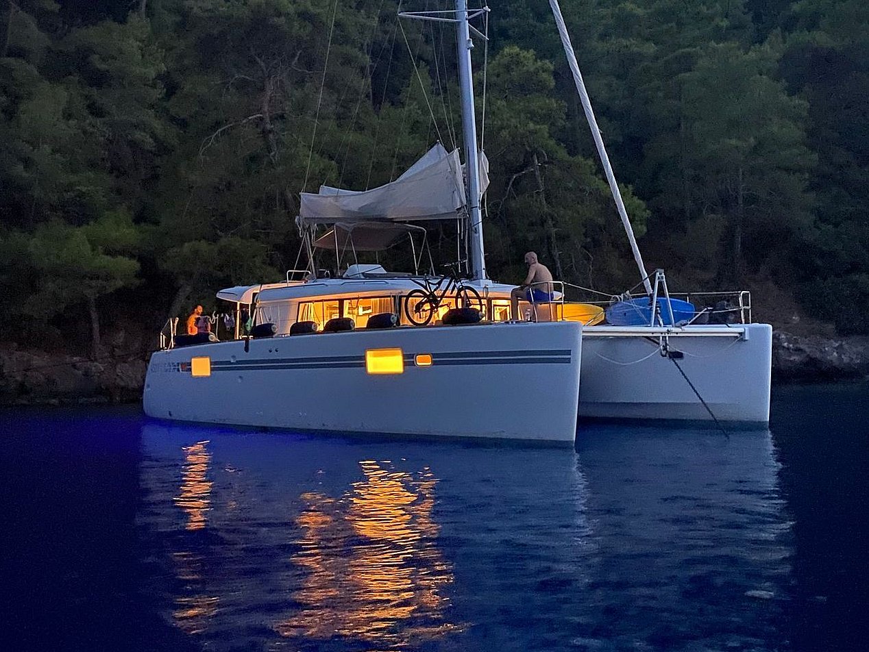 Ağaçların önünde, su üzerinde ışıkları yanan yelkenli katamaran ve güvertede bir kişi.