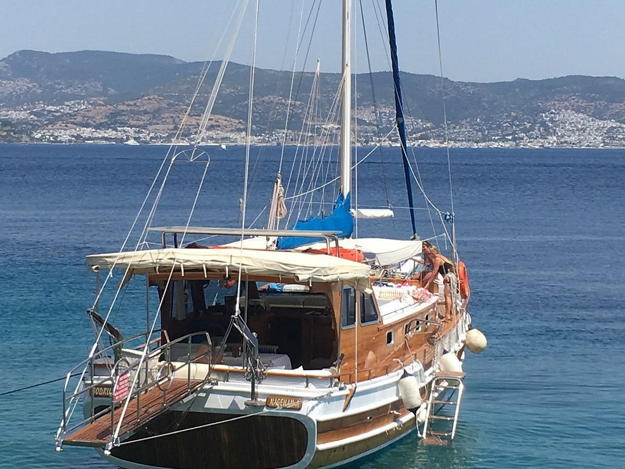 Mavi denizde, Türk bayrağı sallanan ahşap yelkenli tekne ve arka planda dağlar.