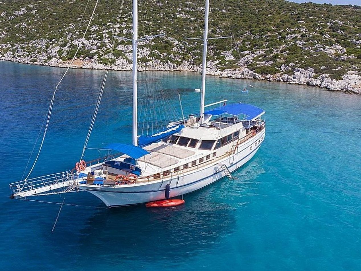 Mavi suda, kıyısında yeşil ve kayalık bir tepenin önünde beyaz yelkenli tekne.