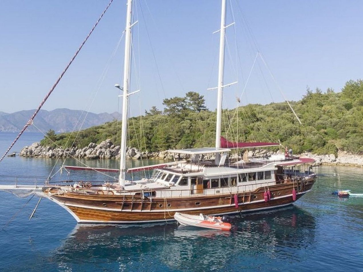 İki direkli, ahşap gövdeli yelkenli tekne, sakin mavi suda, yeşil ağaçların önünde demirli.