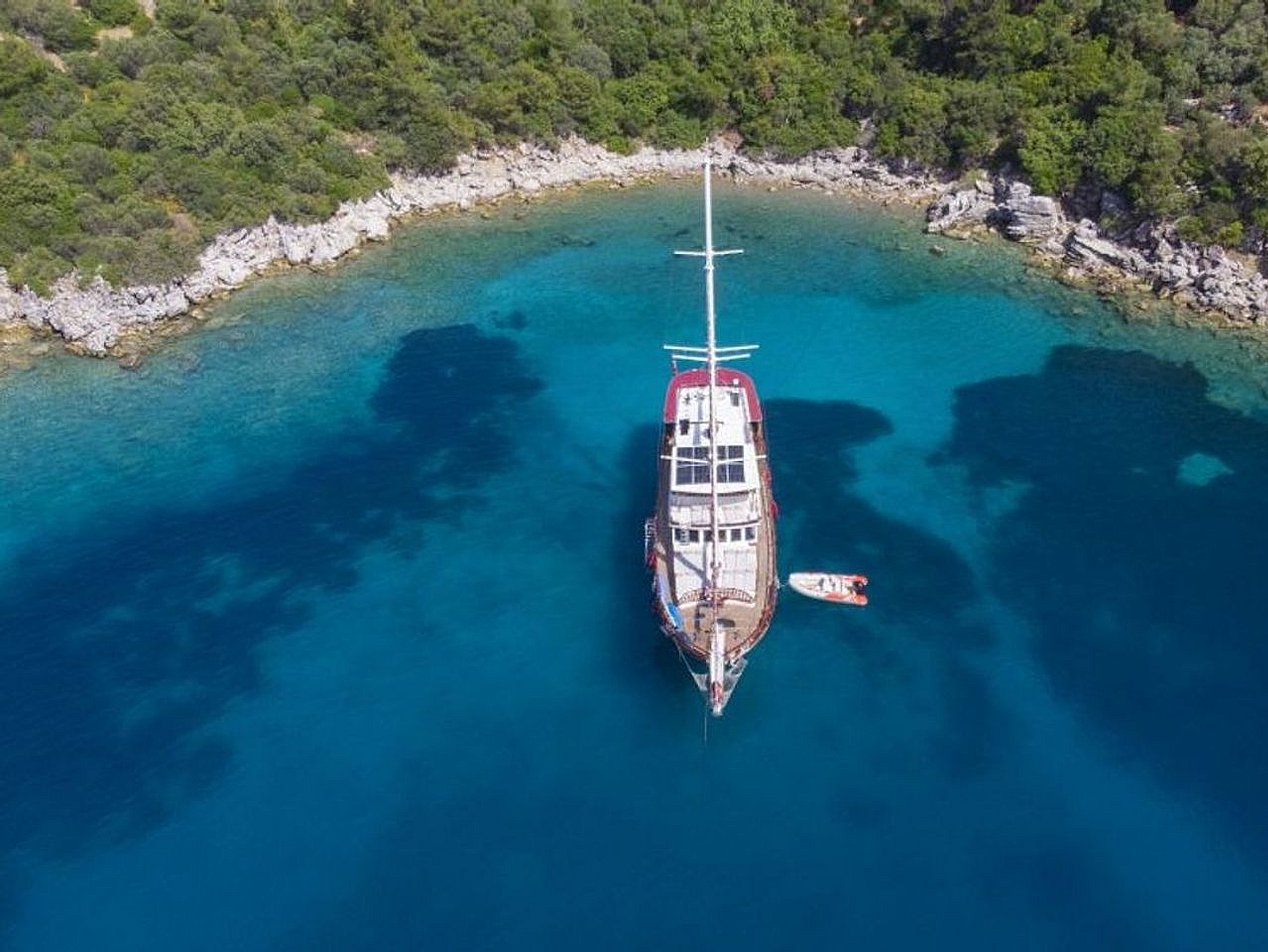 Mavi suda, kıyısında yeşil ağaçlar ve kayalıklar olan yelkenli tekne ve yanında küçük bot.