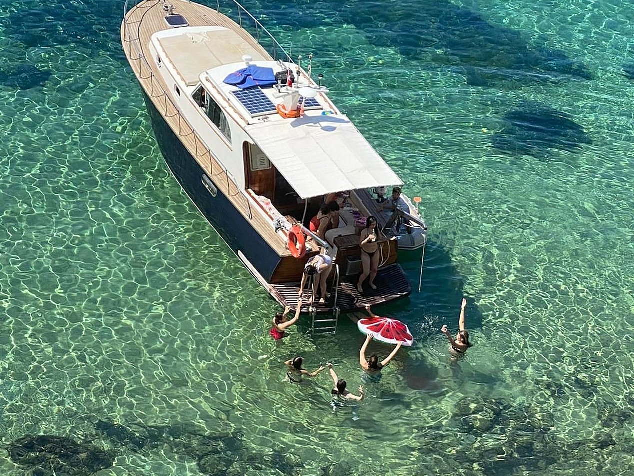 Şeffaf turkuaz suda, kıyıya yakın demirlemiş beyaz ve mavi tekne ve etrafında yüzen birkaç kişi.