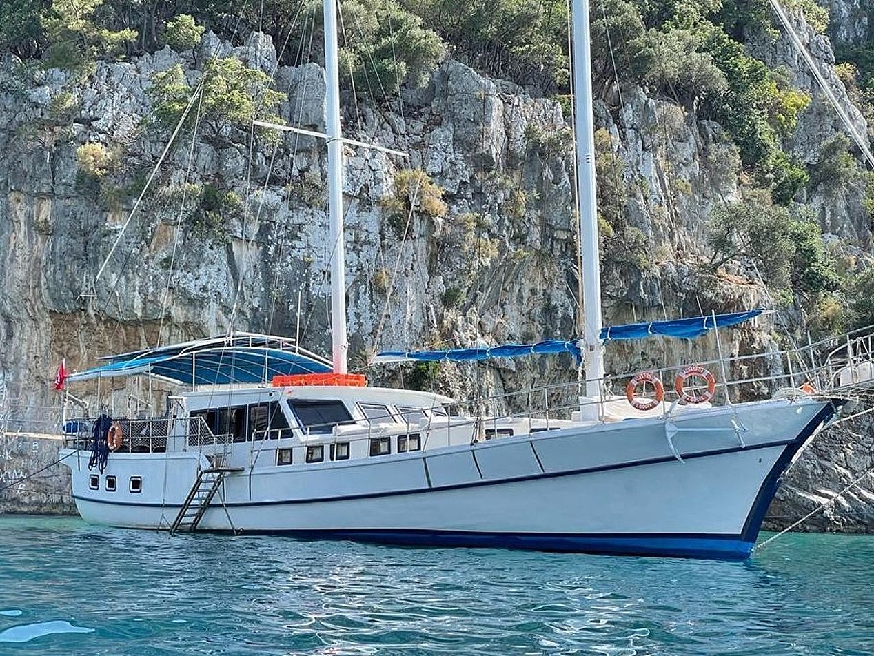 White sailboat on blue water in front of rocky, tree-covered hillside.
