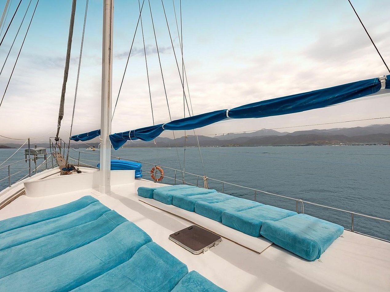 White sailboat with blue cushions on deck and a mast, with sea and mountains in the background.