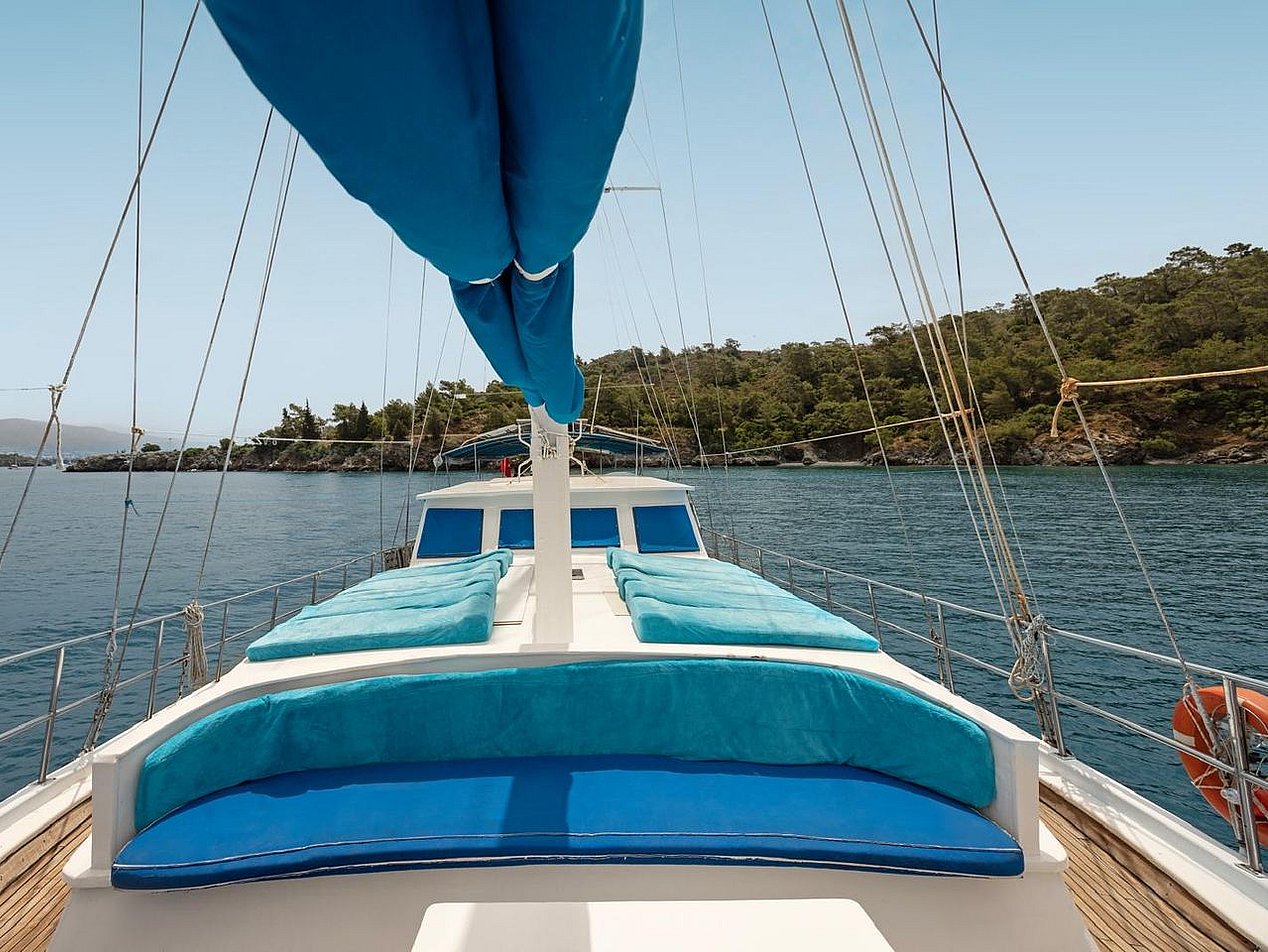 Sailboat deck equipped with blue cushions and covers, sea and tree-lined shore in the background.