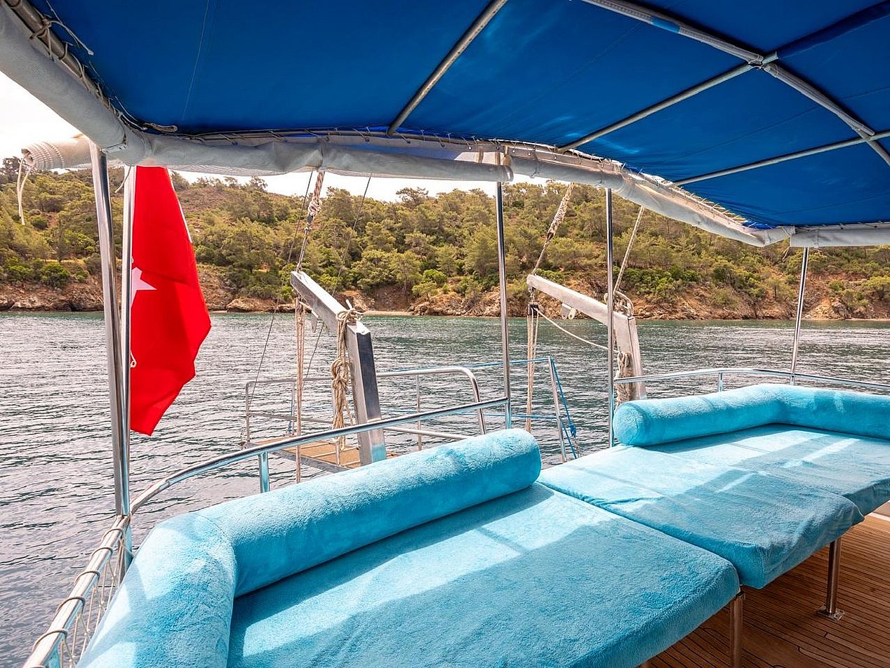 Boat deck with wide seating area covered in blue cushions and a red flag, wooded shoreline in the background.