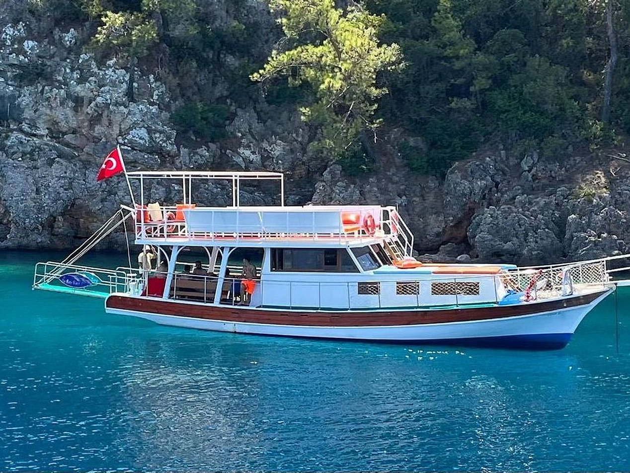 İki Türk bayrağı taşıyan, üst güvertesinde oturma alanı bulunan beyaz ve kırmızı tekne, mavi suda ve kayalık ormanlık alanda.