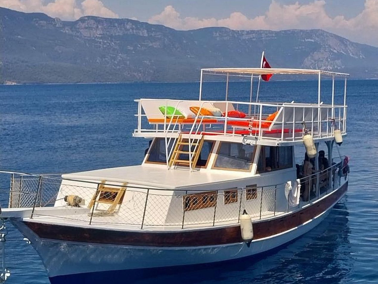 Mavi denizde, üst güvertede renkli minderler ve Türk bayrağı bulunan beyaz ve kahverengi ahşap tekne.