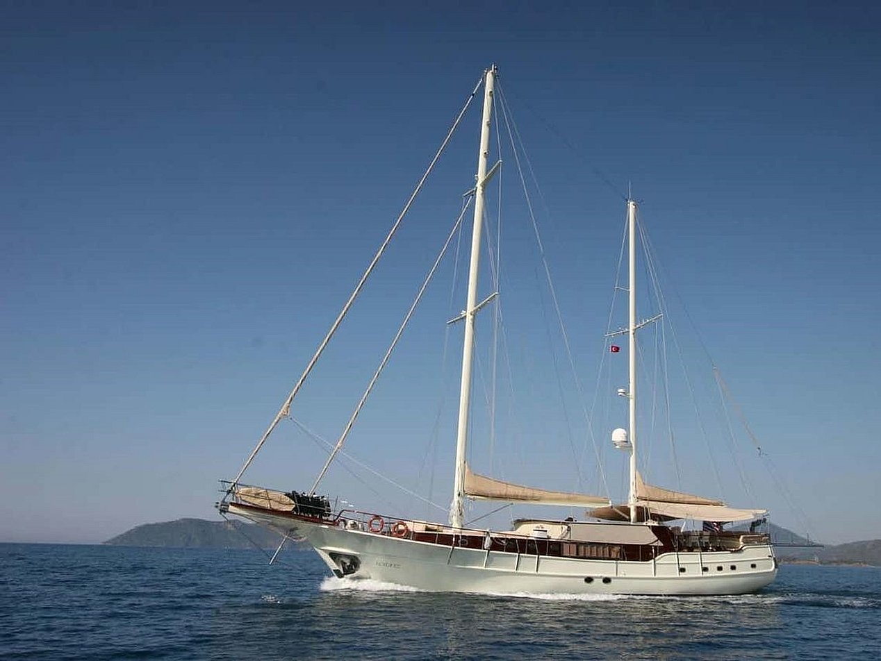 Mavi denizde, iki direkli beyaz yelkenli tekne, arka planda dağlar.