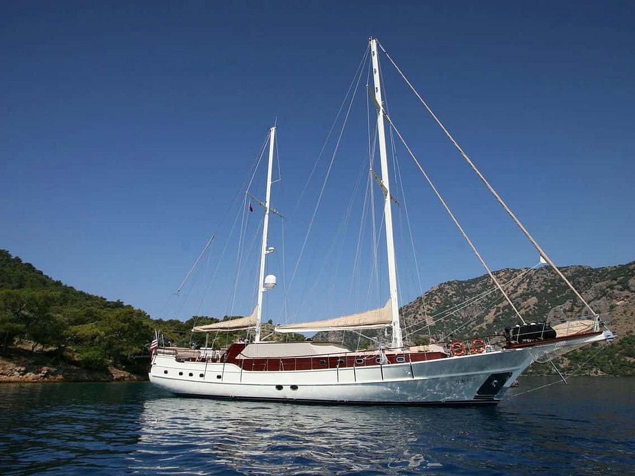 Mavi suda, ormanlık tepelerin önünde beyaz yelkenli tekne.
