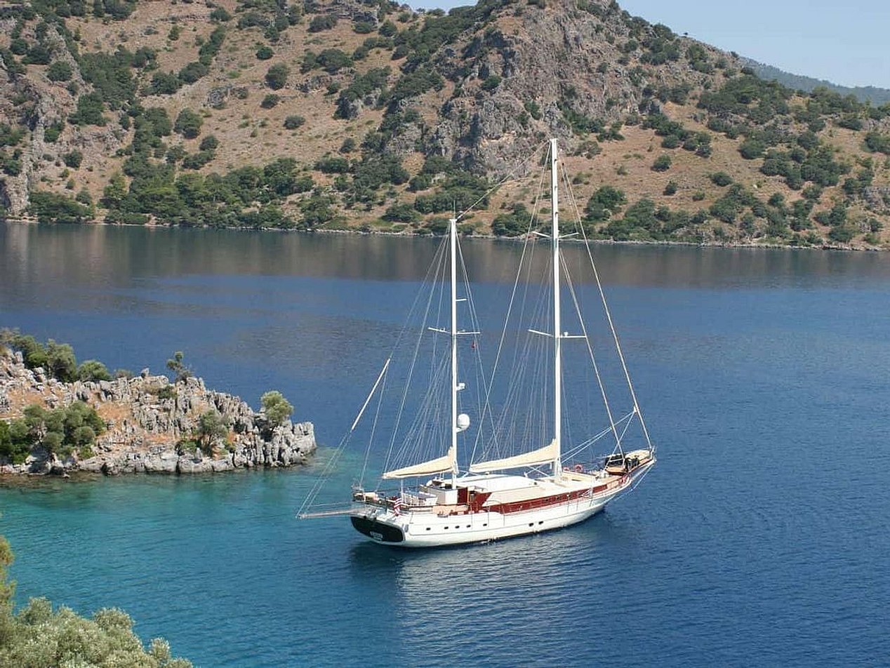 Mavi suda, kıyısında ağaçlı ve kayalık yamaçların olduğu beyaz yelkenli tekne.