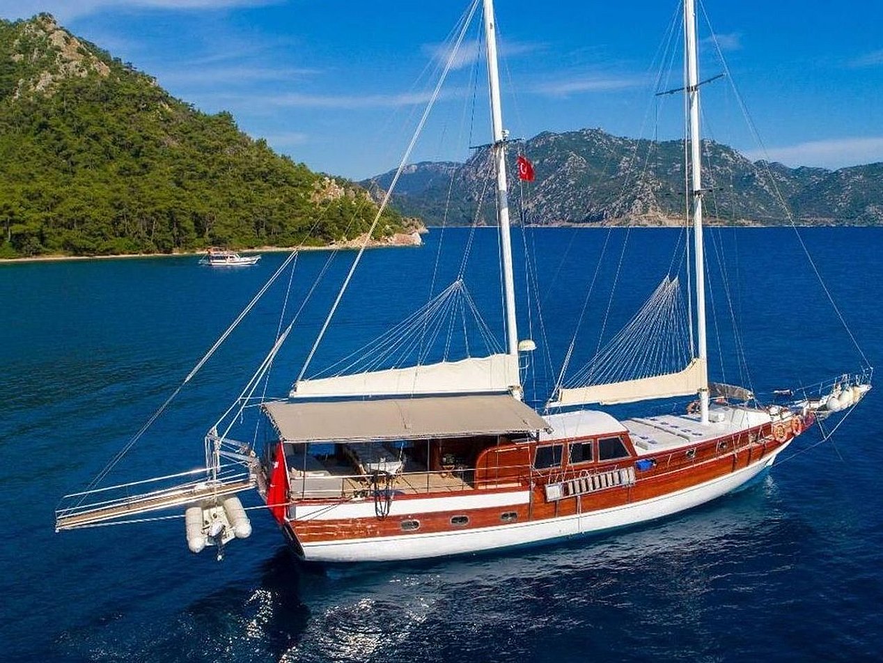 Mavi denizde, ormanlık yeşil tepelerin önünde beyaz ve kahverengi yelkenli tekne.
