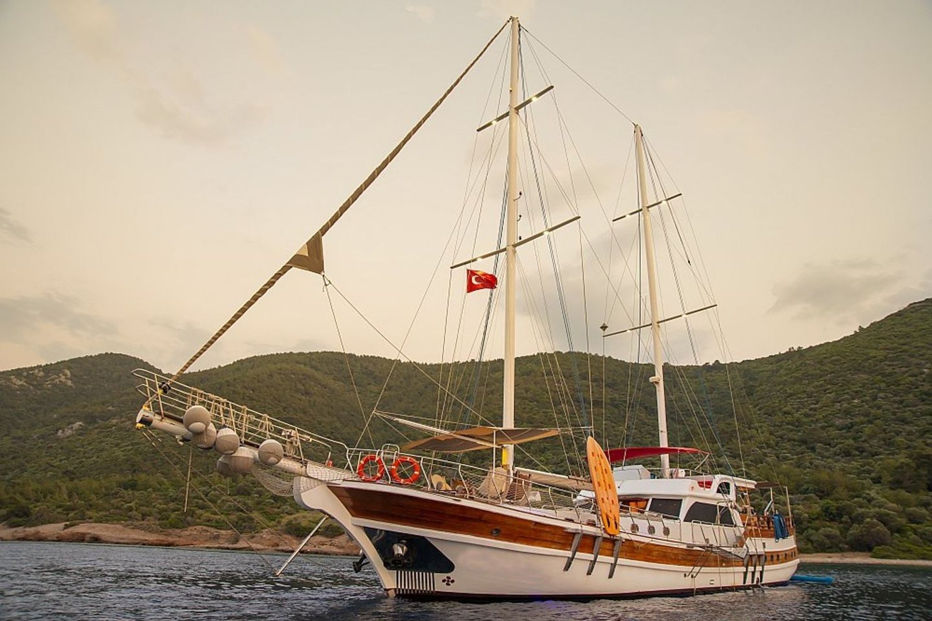 İki direkli, ahşap gövdeli yelkenli tekne, kıyıda yeşil ormanlı dağlar önünde.