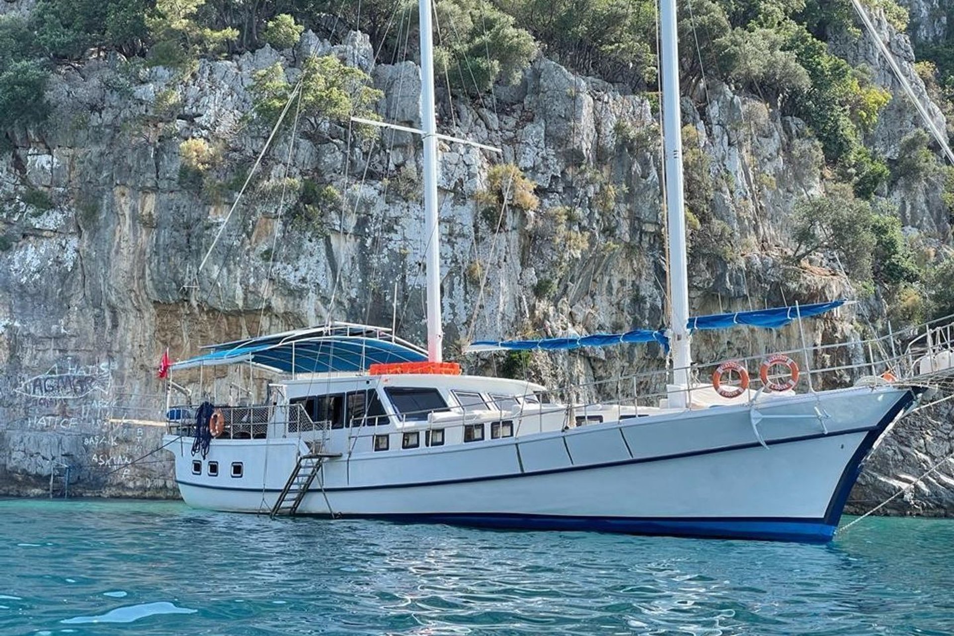 White sailboat on blue water in front of rocky, tree-covered hillside.