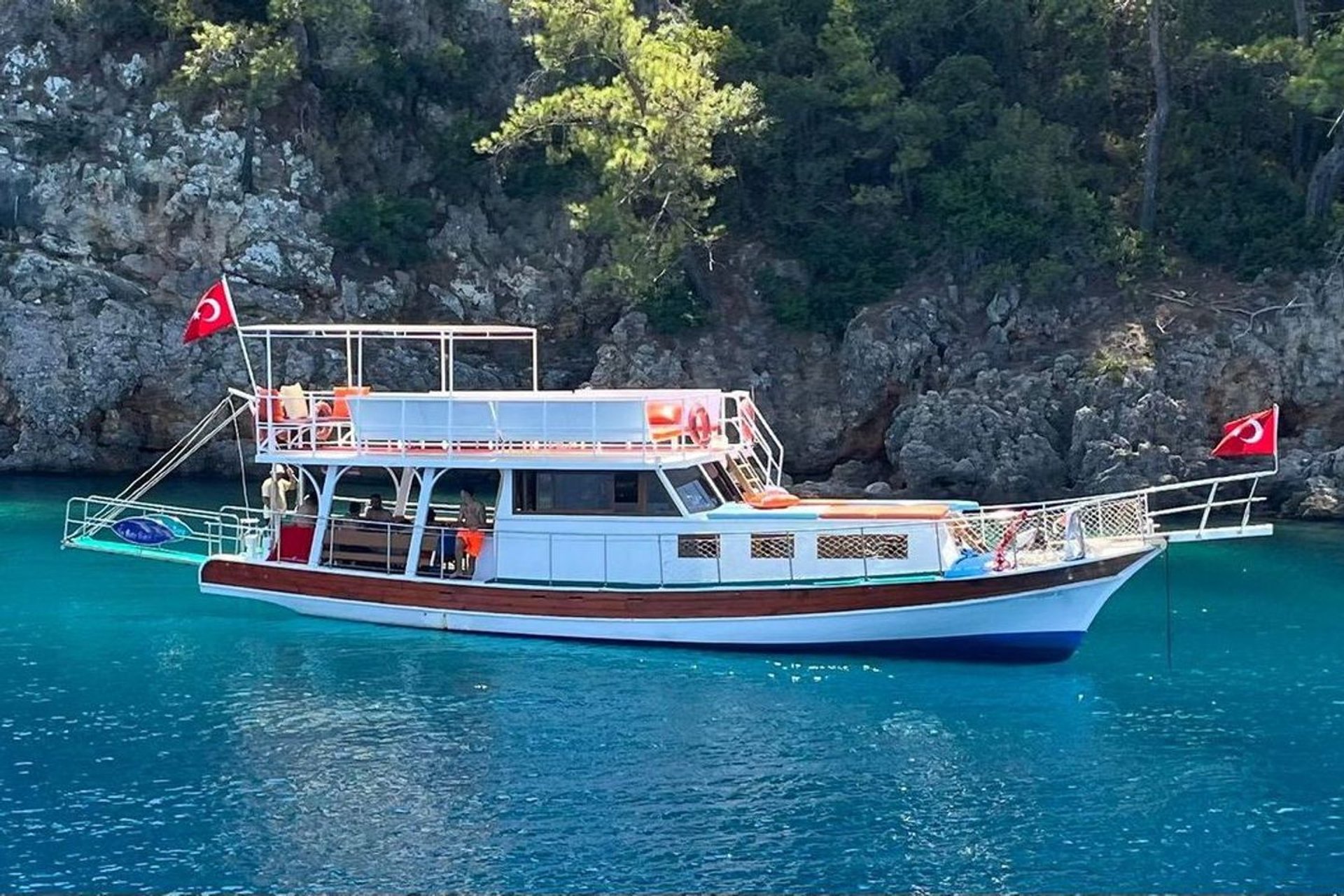İki Türk bayrağı taşıyan, üst güvertesinde oturma alanı bulunan beyaz ve kırmızı tekne, mavi suda ve kayalık ormanlık alanda.