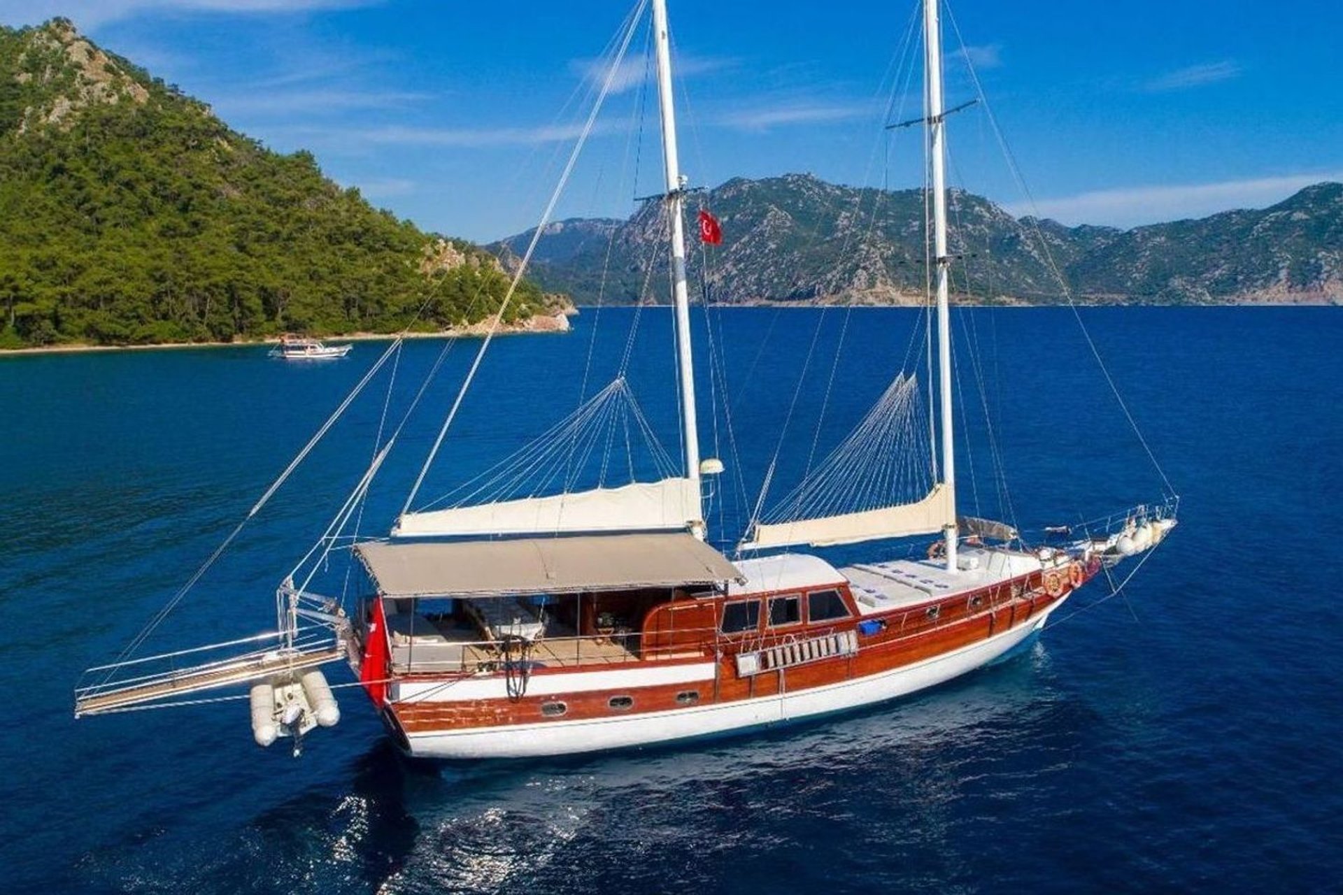 Mavi denizde, ormanlık yeşil tepelerin önünde beyaz ve kahverengi yelkenli tekne.