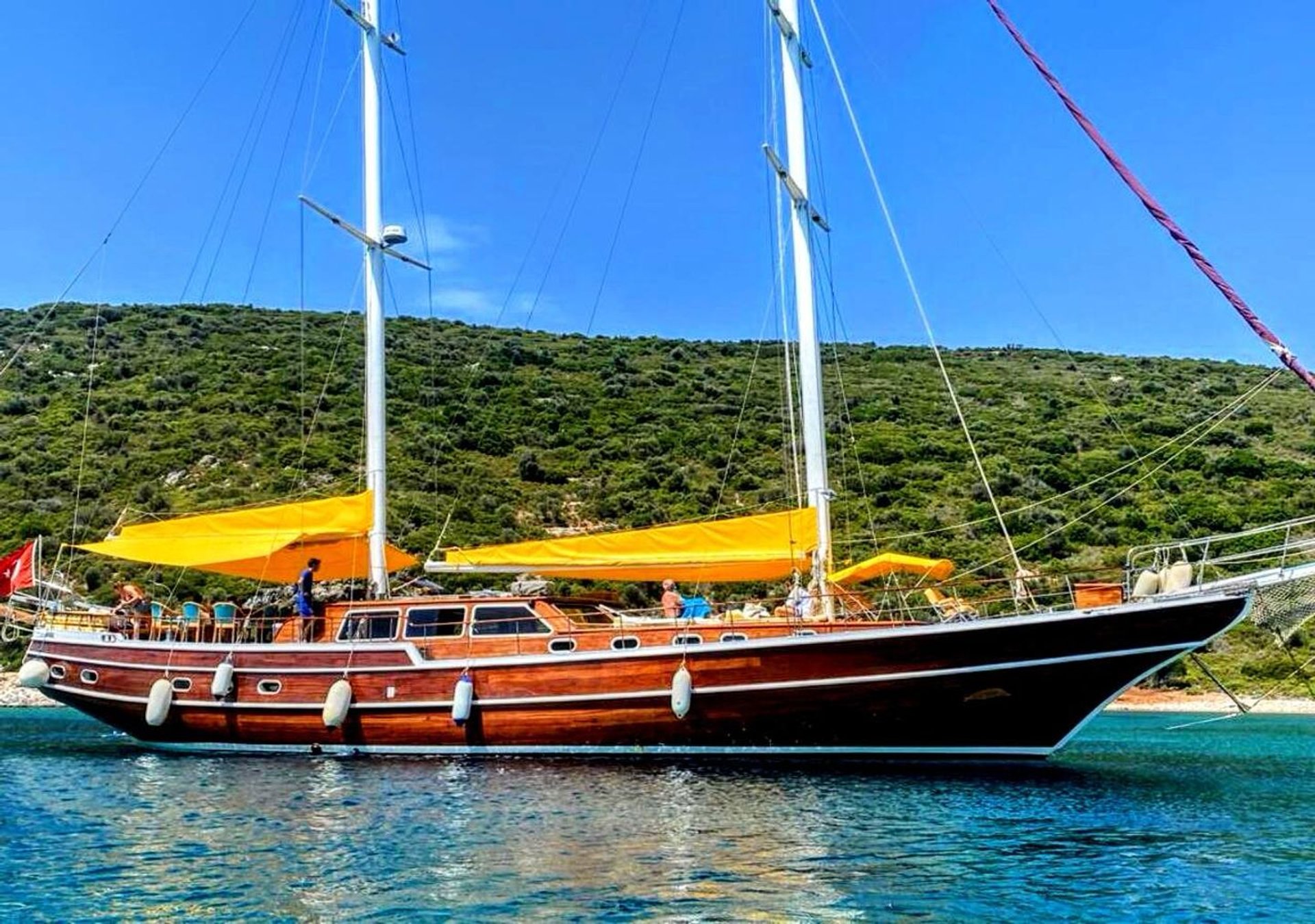 İki direkli, ahşap gövdeli yelkenli tekne, sarı tente ve yeşil tepelerin önünde mavi suda.