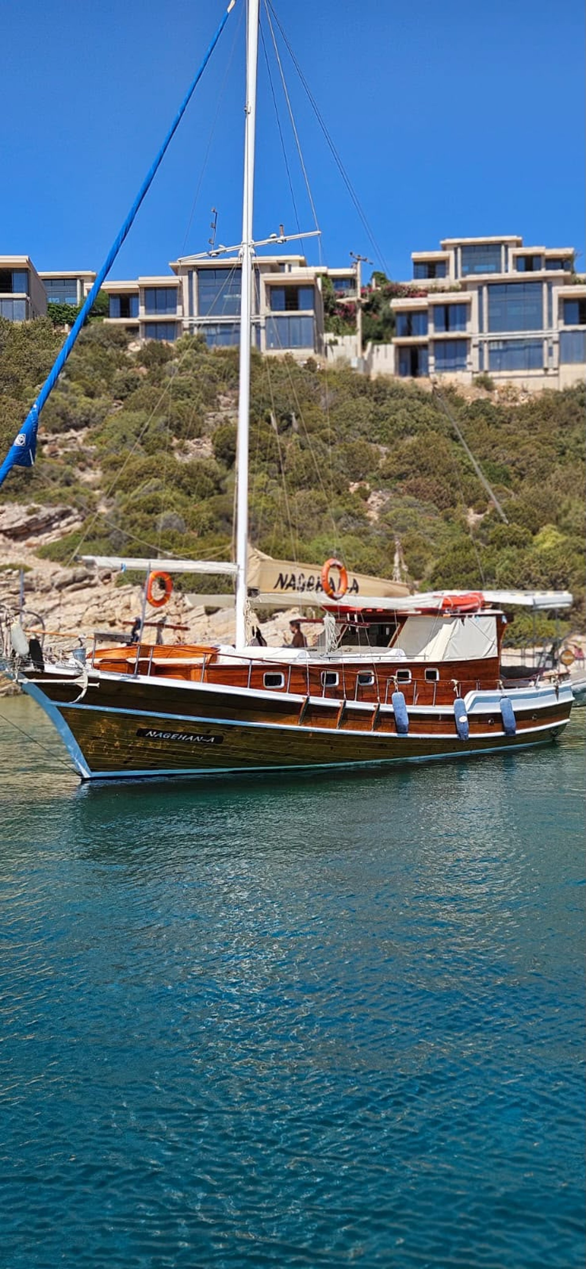 Ahşap gövdeli, beyaz detaylı yelkenli tekne, arka planda yeşil ağaçlar ve modern binalar.